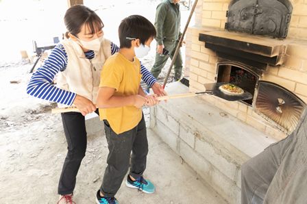 子供が学生に支えられてピザ窯体験を行っている様子